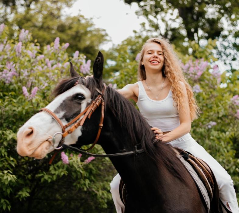 Donna sorridente a cavallo, circondata da fiori e alberi in un ambiente naturale.