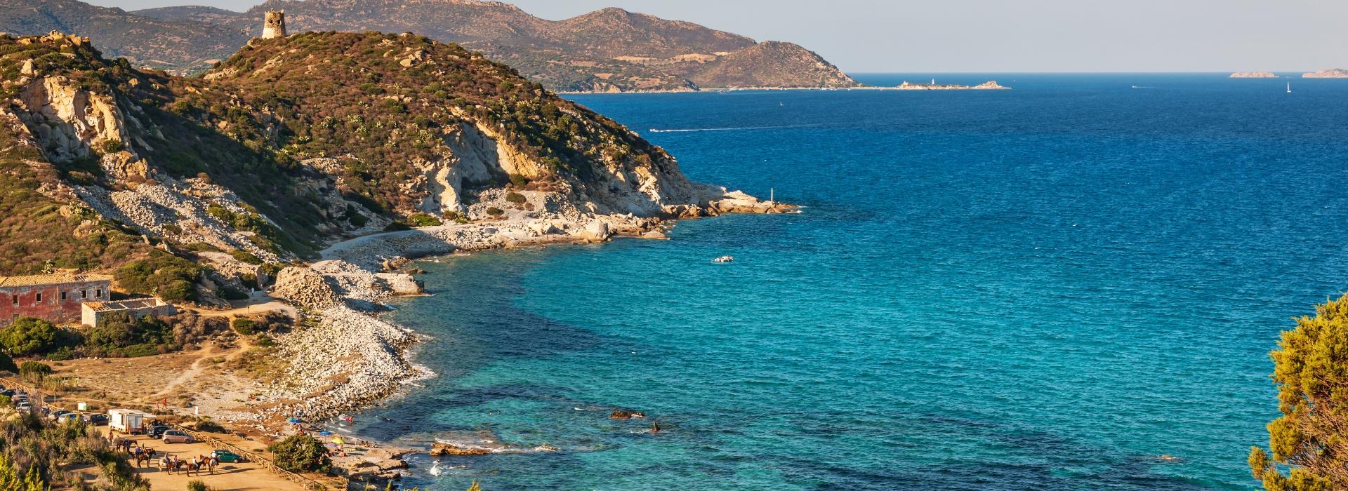 Costa rocciosa con mare cristallino e torre antica in lontananza.