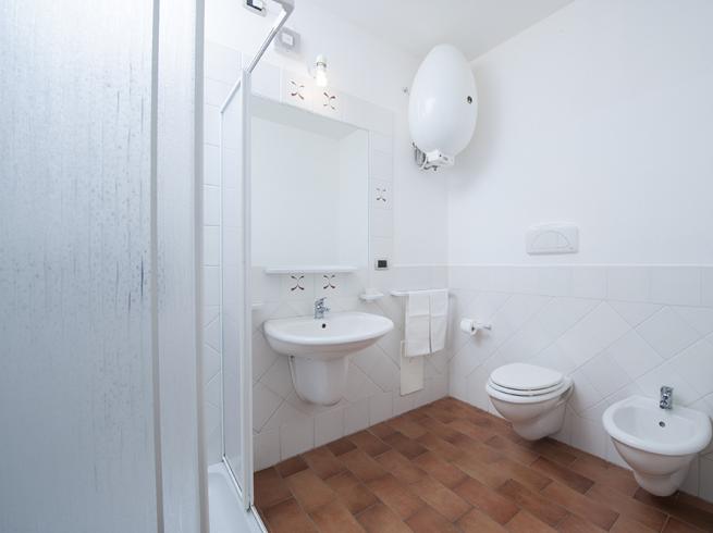 Bagno moderno con doccia, lavabo, WC, bidet e piastrelle bianche su pavimento marrone.